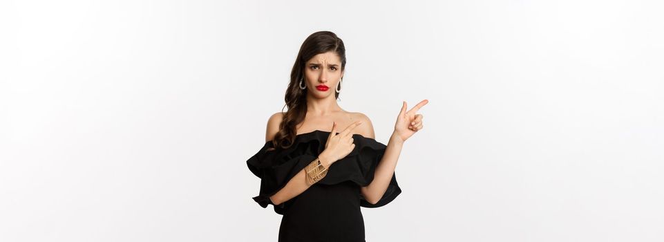 Fashion and beauty. Disappointed glamour woman complaining, wearing black dress, sulking and pointing fingers left at bad offer, white background.