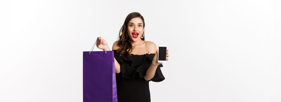 Beauty and shopping concept. Beautiful and stylish woman showing smartphone screen and bag, buying online, standing over white background.