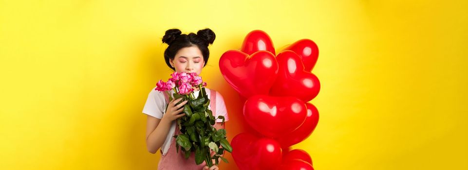 Happy Valentines day. Tender asian girl smelling flower from boyfriend. Girlfriend holding beautiful pink roses, standing near romantic hearts balloons, yellow background.