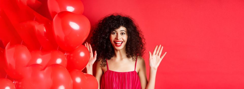 Excited stylish woman with curly hair, wearing dress, raising hands up and laughing happy, standing near heart balloons, white background.