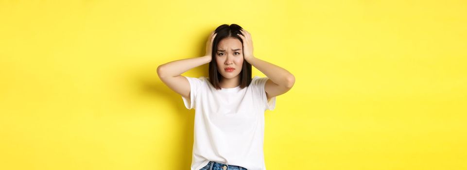 Asian woman holding hands on head and looking sad, having a problem, standing anxious against yellow background.