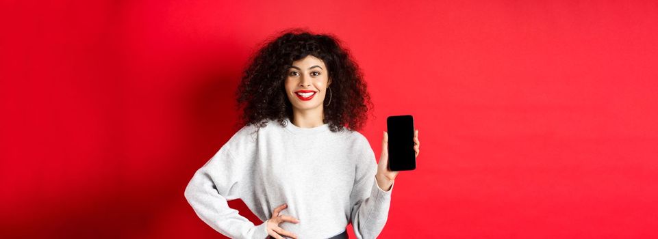 Attractive smiling woman showing empty smartphone screen and looking happy, advertising online store or application, standing against red background.