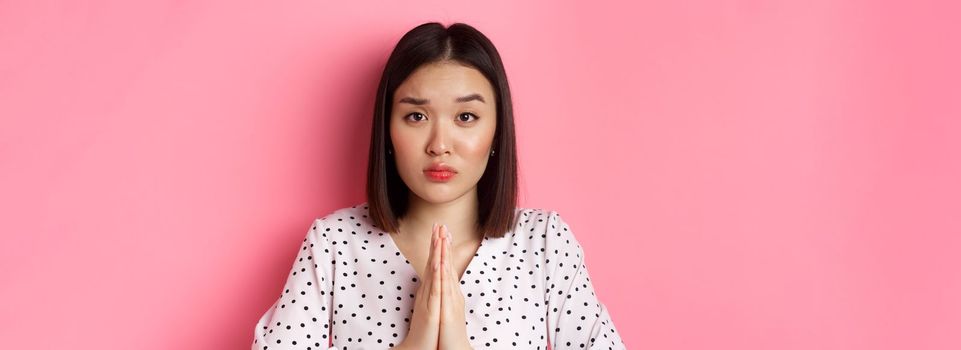 Beauty and lifestyle concept. Sad asian woman asking for help, begging with hands in plead gesture, staring at camera, need favour, standing over pink background.