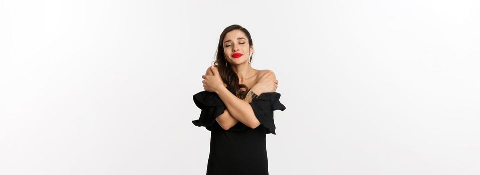Fashion and beauty. Sensual and beautiful woman in black dress, hugging herself with closed eyes, daydreaming, standing over white background.