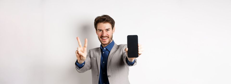 E-commerce and online shopping concept. Cheerful man in business suit showing v-sign and empty mobile phone screen, demonstrate smartphone app, white background.