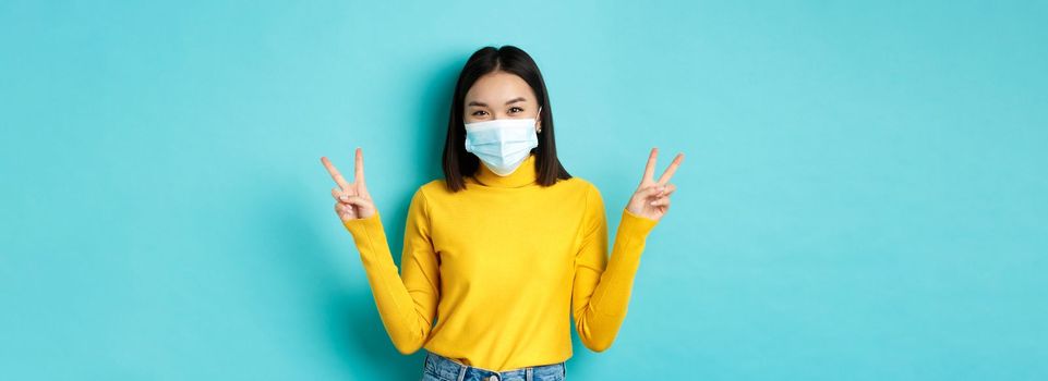 Covid-19, social distancing and pandemic concept. Kawaii asian woman showing peace signs and smiling happy, wearing medical mask from coronavirus disease, standing over blue background.
