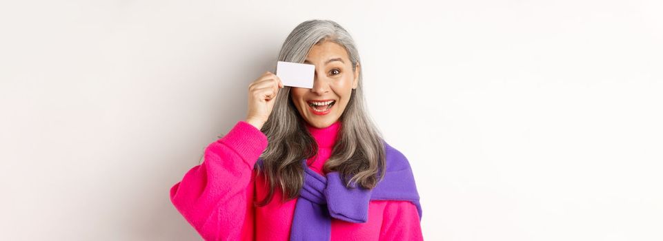 Shopping concept. Stylish asian senior woman smiling and showing plastic credit card, paying contactless, standing over white background.