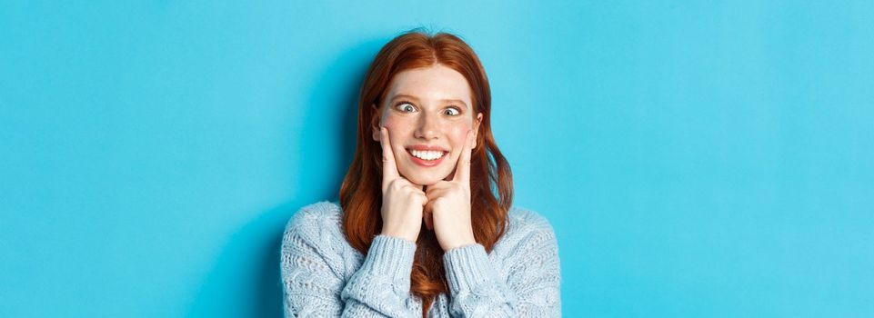 Close-up of funny redhead teen girl making faces, squinting and squeezing cheeks, standing against blue background.