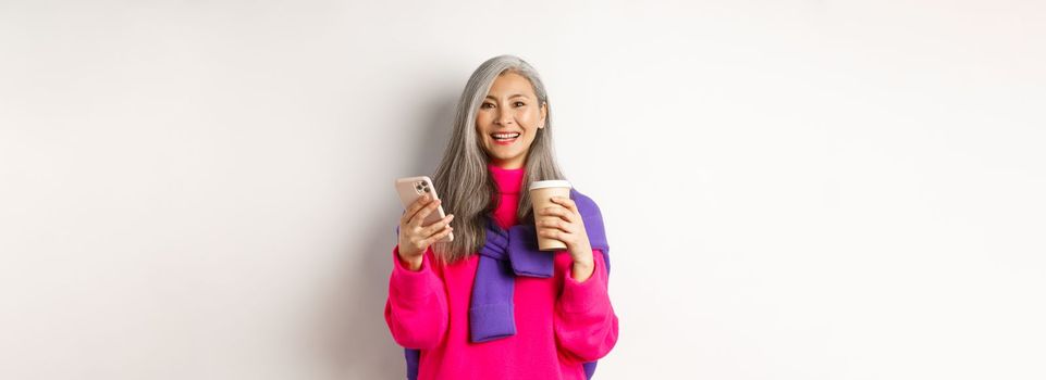 E-commerce concept. Beautiful asian senior woman drinking coffee from takeaway cup and using mobile phone, smiling at camera, white background.