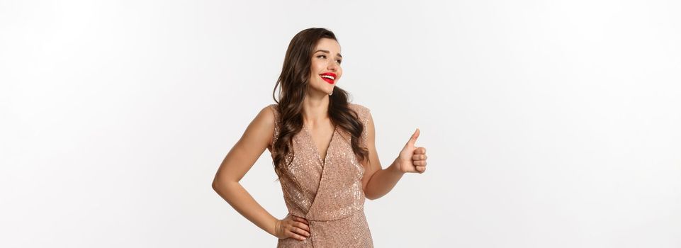Concept of celebration, holidays and party. Beautiful female model in luxurious dress, looking left and showing thumb-up in approval, praise good choice, standing over white background.
