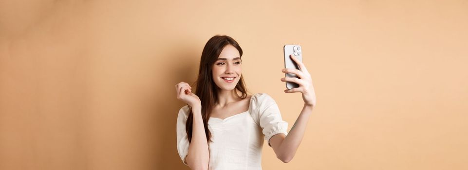 Beautiful woman taking selfie on smartphone, posing for phone photo, standing on beige background.