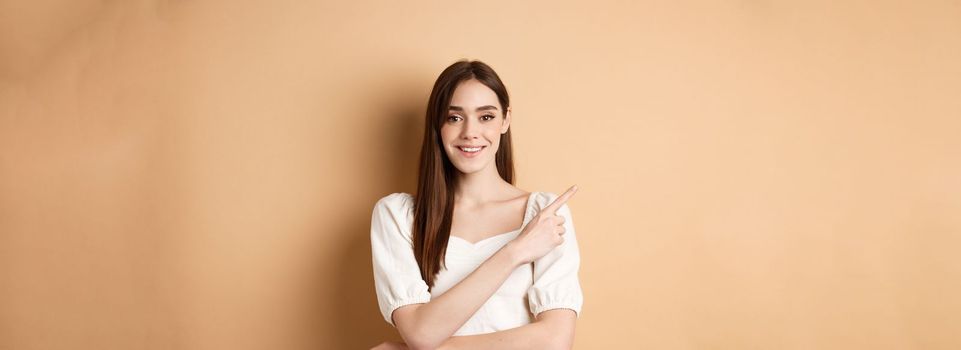 Young cheerful female model in romantic dress pointing finger at upper left corner, smiling pleased at camera, showing her choice, recommending product, beige background.