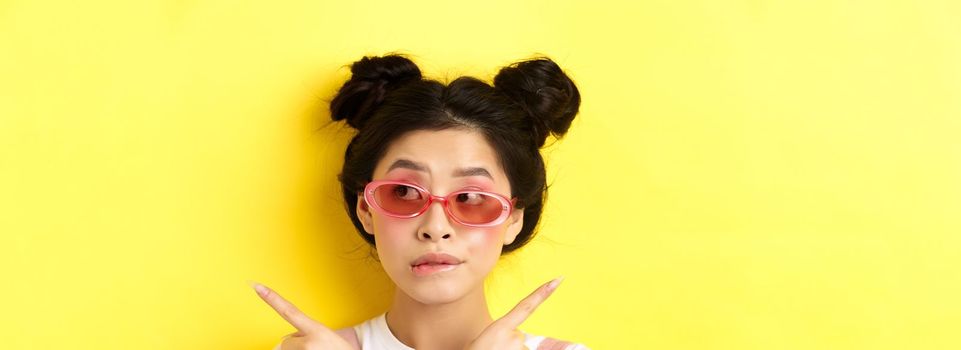 Summer fashion concept. Pensive asian girl making choice, pointing sideways and looking thoughtful, deciding what pick, standing in sunglasses on yellow background.