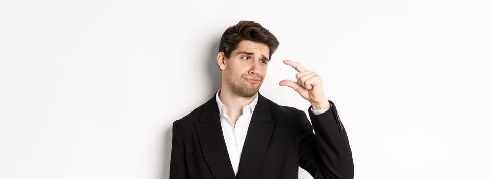 Close-up of handsome businessman in trendy suit, showing something small with disappointment, standing against white background.