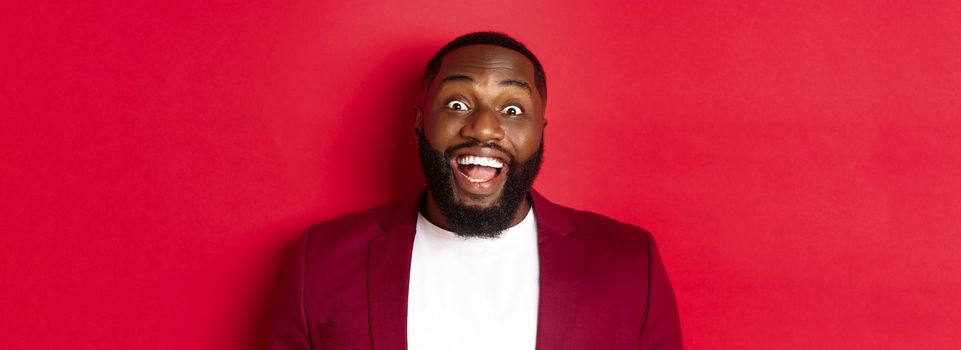Close-up of african american man with beard staring amazed at camera, smiling and looking excited, standing in blazer against red background.