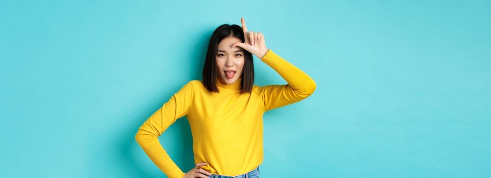 Asian girl winner mocking over lost team, showing loser sign on forehead and sticking tongue ignorant, standing over blue background.