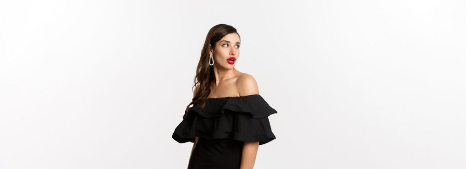 Fashion and beauty concept. Image of attractive young woman in black dress turn behind and looking at copy space, standing over white background.