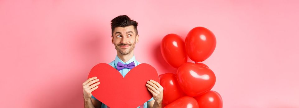Valentines day concept. Smiling handsome man looking dreamy left, waiting for true love girlfriend with big red heart cutout, standing over pink background.