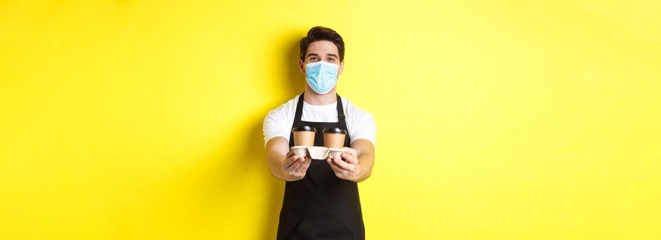 Concept of covid-19, cafe and social distancing. Barista in medical mask serving coffee in takeaway cups, standing in black apron against yellow background.