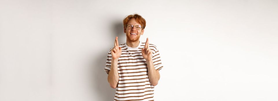 Happy redhead man with bristle cross fingers for good luck and smiling hopeful, making wish, standing over white background.