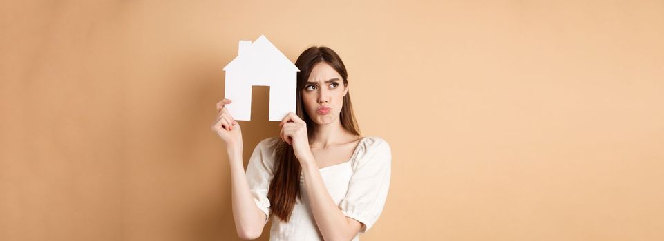 Real estate. Upset young woman showing paper house cutout and frowning sad, looking aside thoughtful, thinking of buying apartment, standing on beige background.