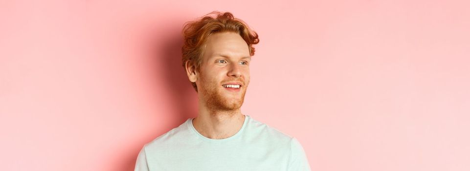 Portrait of attractive caucasian guy with red messy hair and beard, turn head and looking left with pleased smile, standing over pink background.