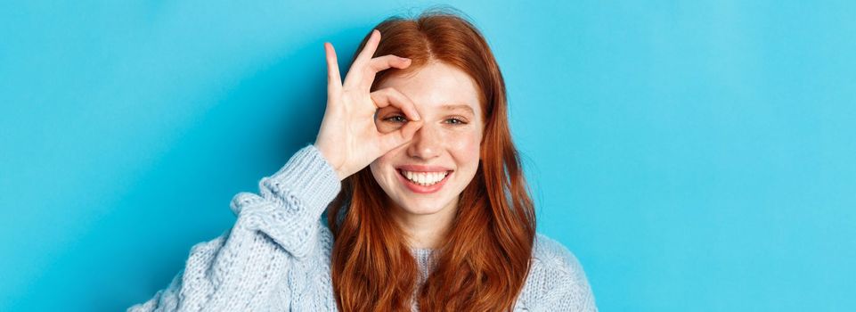 Headshot of cheerful redhead female model showing okay sign over eye, smiling satisfied and happy, standing against blue background.