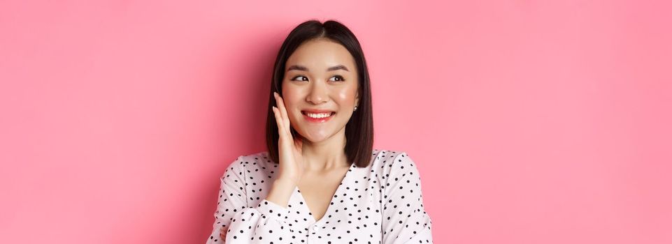 Coquettish asian woman in blushing, touching cheek and looking left amused, standing over pink background.