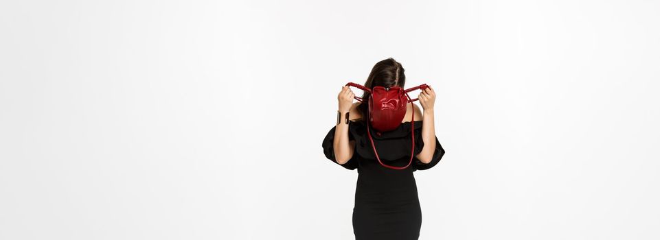 Beauty and fashion concept. Full length of young woman sticking head inside purse and searching something, wearing black dress and high heels, standing over white background.