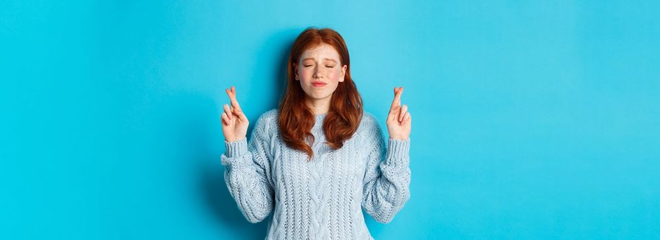 Hopeful redhead girl making a wish, cross fingers for good luck, smiling and anticipating good news or positive result, standing against blue background.