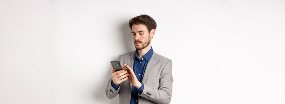 Young businessman in suit texting message on mobile phone, looking at smartphone, standing against white background.