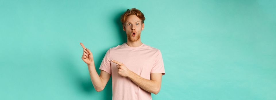 Handsome caucasian man with red hair, gasping in awe, pointing fingers at upper right corner, checking out promotion deal, standing over turquoise background.