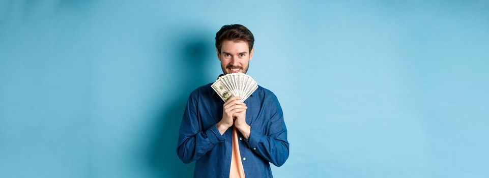 Happy smiling man holding money and looking at camera pensive, thinking of shopping, standing on blue background.