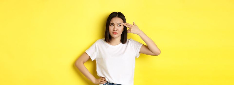 Woman showing kill me please gesture, shooting herself in head with finger gun from boredom, standing upset over yellow background.
