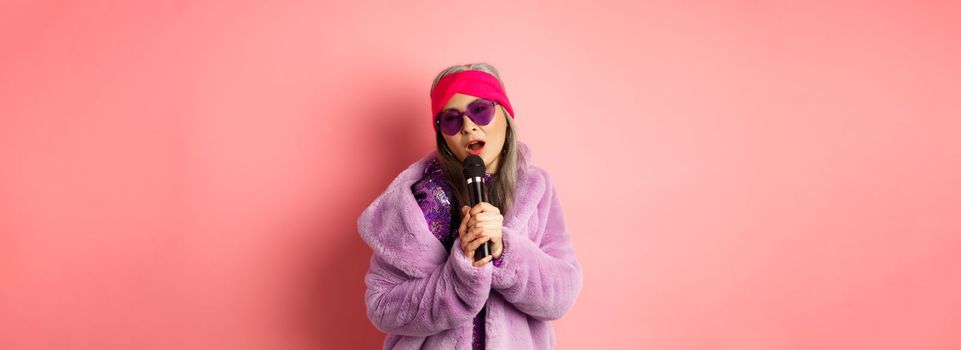 Fashion concept. Stylish senior asian woman singing karaoke, perform on stage with microphone, wearing trendy sunglasses and purple faux fur coat, pink background.