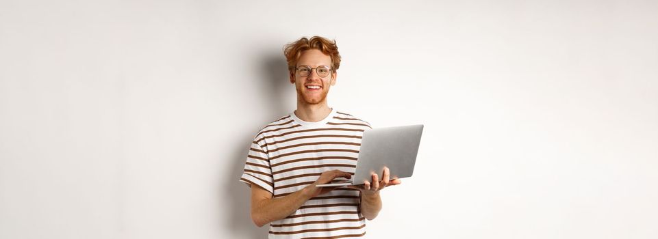 Smiling young man working on laptop and looking joyful, standing over white background.