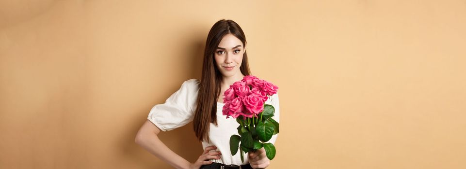 Valentines day. Beautiful girlfriend holding pink roses and looking at camera. Young woman receive flowers from her date, standing on beige background.