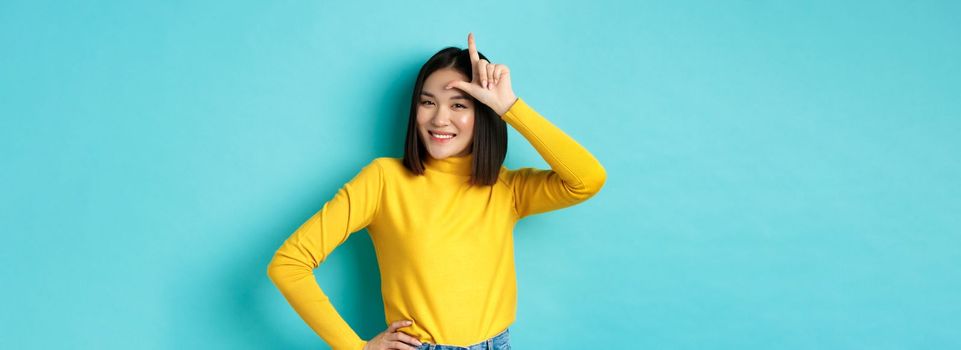 Sassy asian girl mocking lost team, showing loser sign on forehead and smiling pleased, being a winner, standing over blue background.