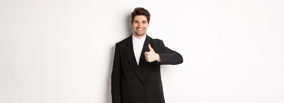 Portrait of handsome and confident male realtor, showing thumb-up and smiling, guarantee quality and recommending company, standing over white background.