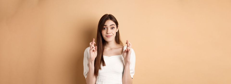 Hopeful tender girl in white blouse making wish, holding fingers crossed and smiling, praying for good luck, waiting for results, beige background.