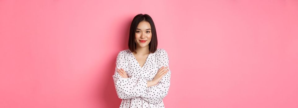 Beautiful asian female model standing in dress, cross arms on chest and smiling at camera, standing over pink background.