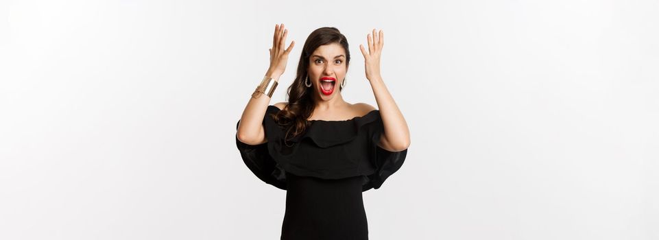 Fashion and beauty. Angry woman in black dress, shouting mad and shaking hands, grimacing outraged at camera, standing over white background.