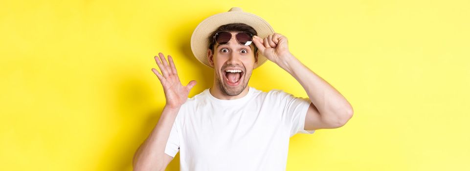 Concept of tourism and holidays. Excited man tourist take-off sunglasses and looking amazed at promo offer, standing over yellow background.