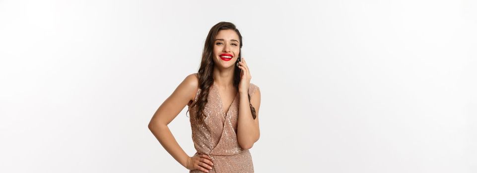 Christmas party and celebration concept. Attractive young woman with red lips, wearing luxury dress, talking on mobile phone and smiling, standing over white background.