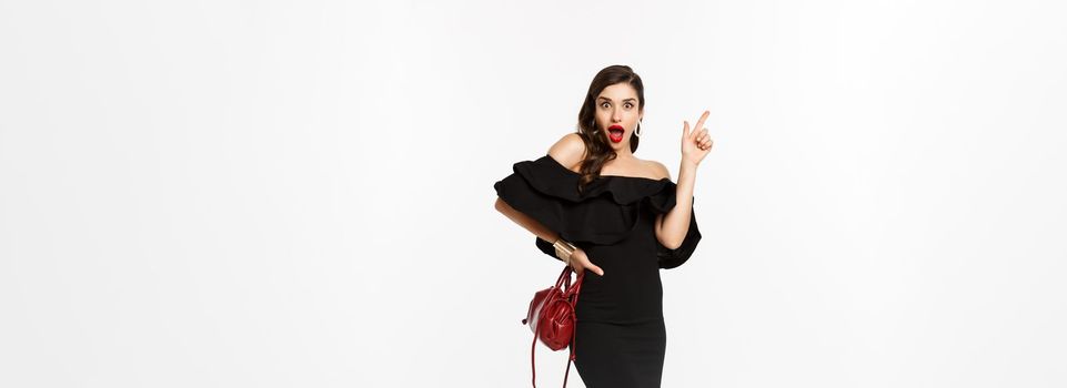 Beauty and fashion concept. Full length of excited young woman in glamour dress, red lips, having an idea, raising finger to suggest something, white background.