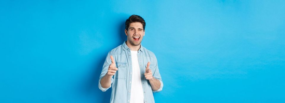 Confident adult man winking, pointing finger at you and winking, congratulating or praising person, standing against blue background.