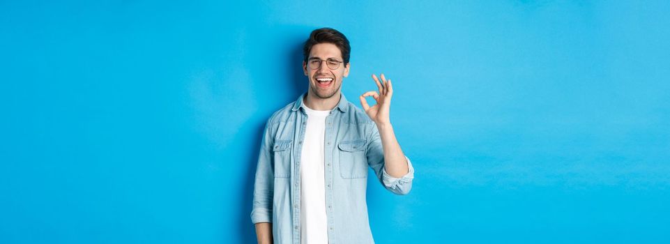 Confident smiling man in glasses showing ok sign, winking to guarantee or recommend something, blue background.