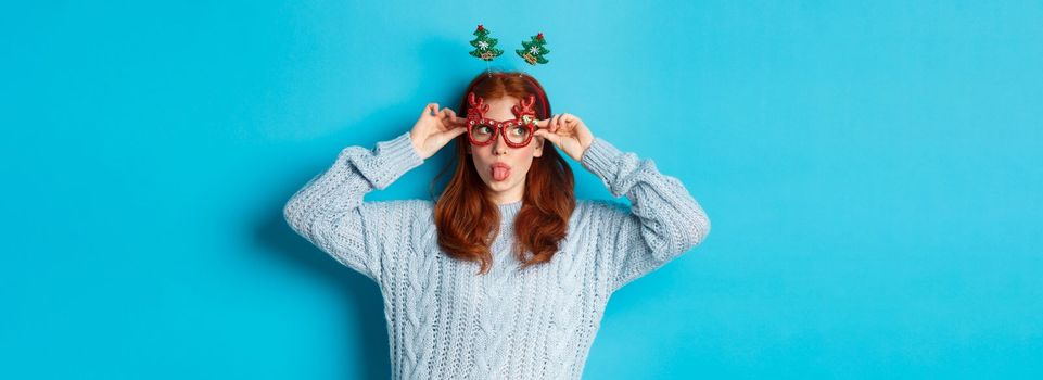 Christmas party and celebration concept. Silly redhead girl enjoying New Year, wearing funny glasses and headband, showing tongue and staring left at logo, blue background.