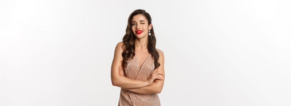 Beautiful woman in evening dress and red lipstick, winking at camera and smiling, celebrating New Year party, standing over white background.