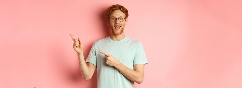 Amazed redhead man in glasses and t-shirt showing special promotion, pointing fingers at upper right corner and staring excited at camera, standing over pink background.